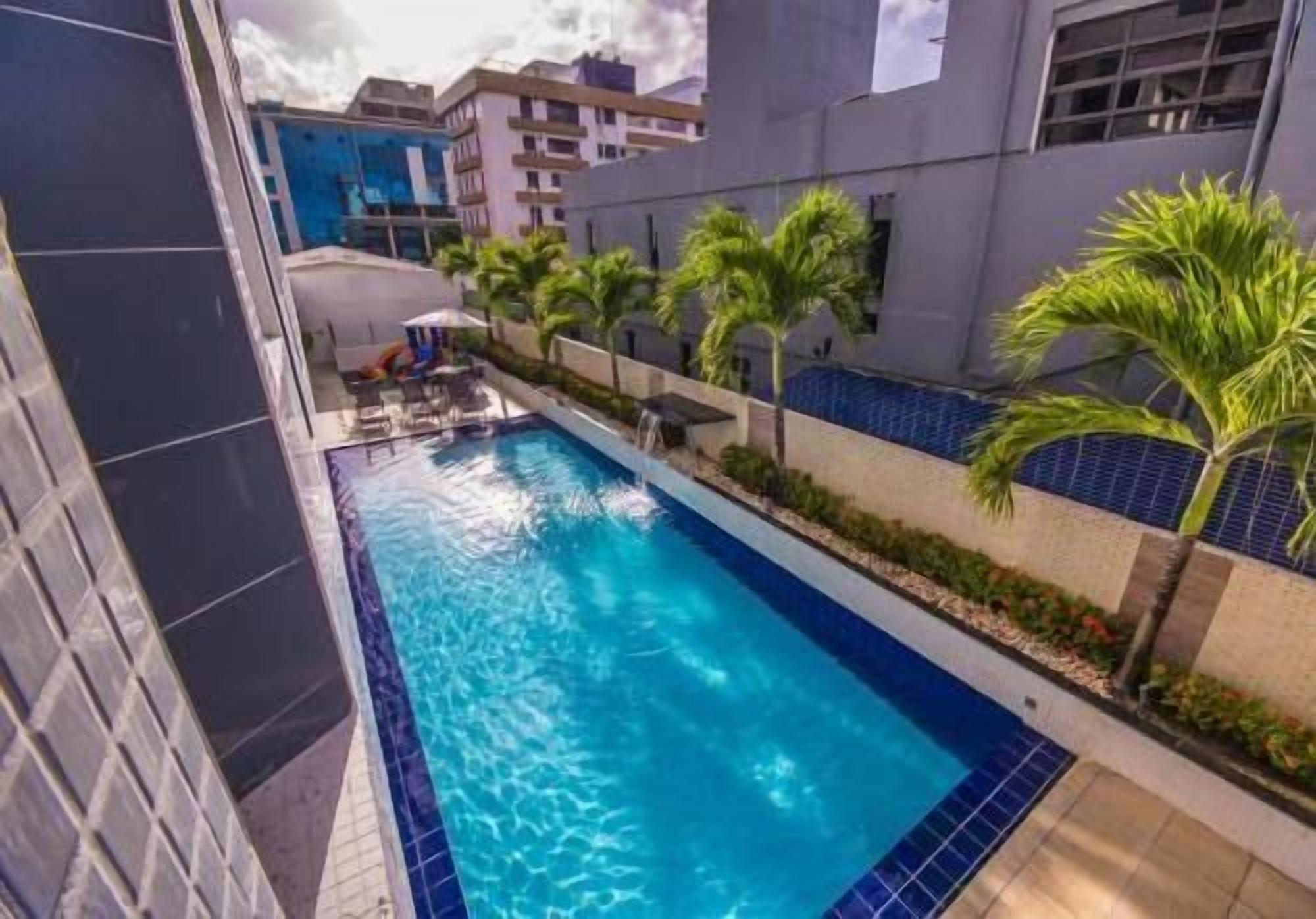A swimming pool with lounge chairs, palm trees, and modern buildings in the background.
