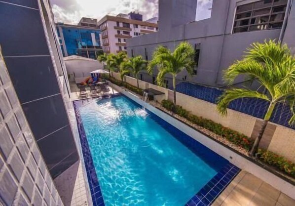 A swimming pool with lounge chairs, palm trees, and modern buildings in the background. - Imperial Suítes (João Pessoa)