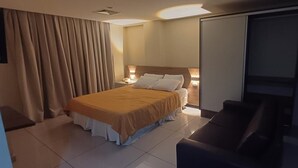 A hotel room with a bed, a desk, a chair, a sliding door, and a nightstand with a lamp. - Imperial Suítes (João Pessoa)