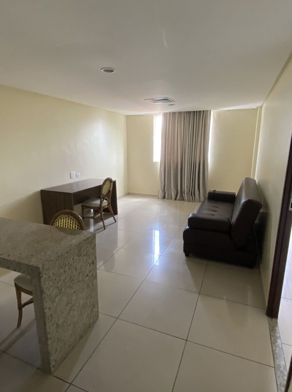 A living room with a sofa, a dining table with chairs, a window with curtains, and a tiled floor.