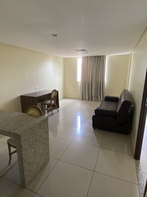 A living room with a sofa, a dining table with chairs, a window with curtains, and a tiled floor. - Imperial Suítes (João Pessoa)