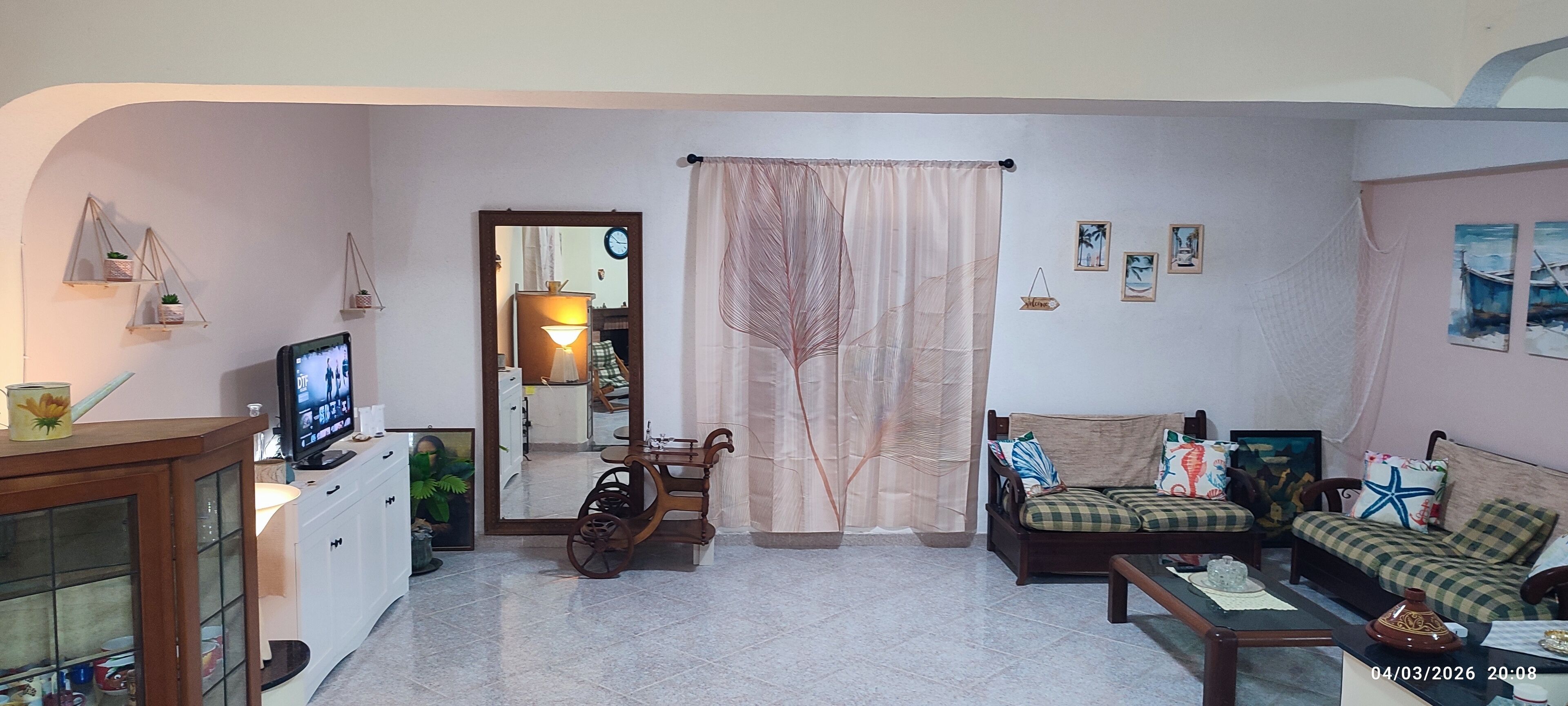 A living room with a sofa, a coffee table, a television, and a cabinet. There is a mirror on the wall and a window with curtains.
