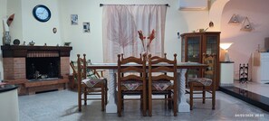A dining area with a wooden table and chairs, a fireplace, a clock, and a window with sheer curtains.