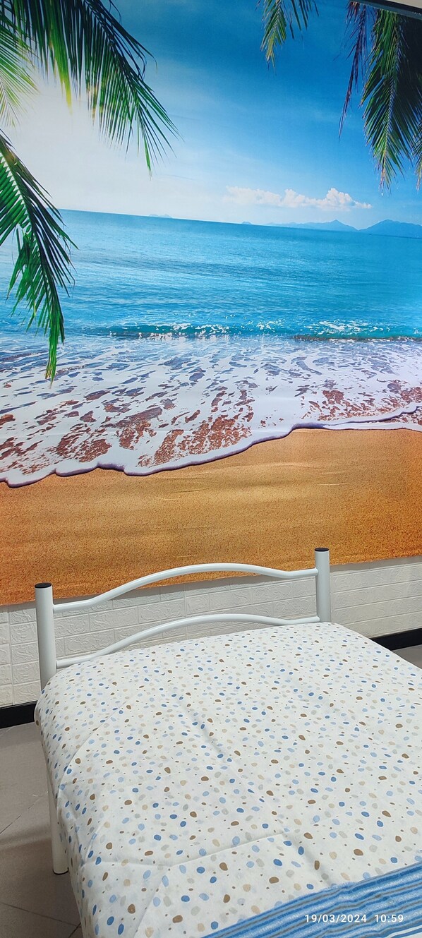 Una cama con estructura blanca y colcha estampada, una pared blanca y vistas a una playa con palmeras y el océano.