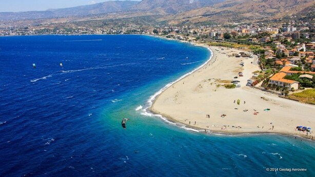 An aerial view of a coastal town with a beach, clear blue waters, and mountainous terrain in the background.