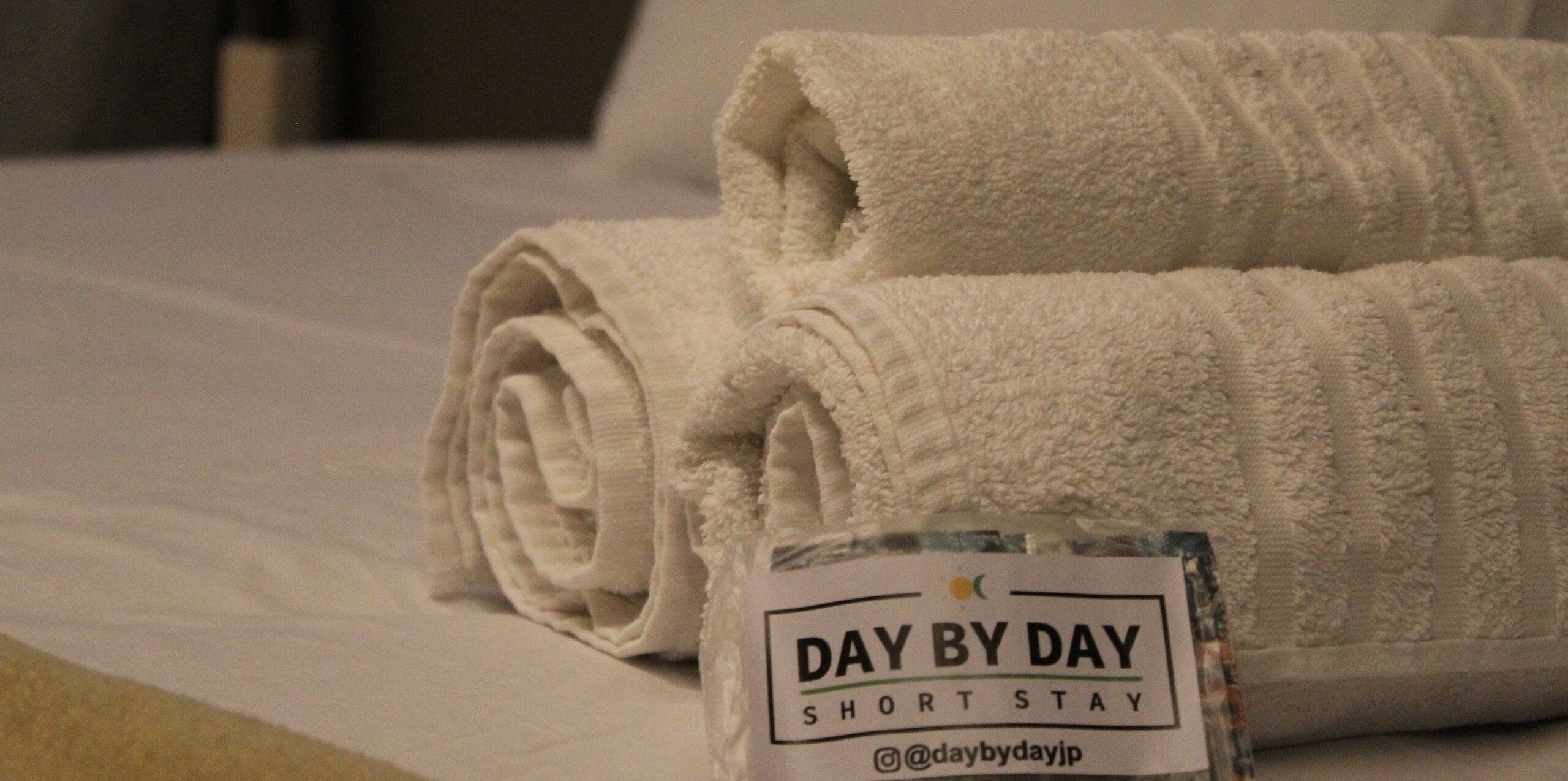 Una cama bien hecha con toallas blancas dobladas y un letrero que dice 'DAY BY DAY SHORT STAY'.