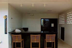 A modern kitchen with a black countertop, three wicker chairs, and a sign reading "Area Gourmet". - Compact Flat Tambau (João Pessoa)