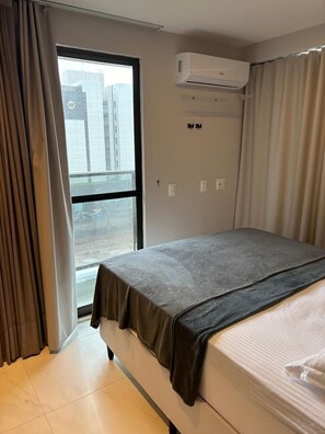 A hotel room with a bed, an air conditioning unit, a window with curtains, and a view of buildings outside. - Compact Flat Tambau (João Pessoa)