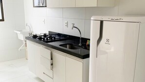 A modern kitchen with white cabinetry, a black countertop, a stainless steel sink, and a white refrigerator. - Compact Flat Tambau (João Pessoa)