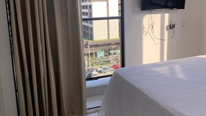 A hotel room with a bed, a TV mounted on the wall, and a view of buildings outside. - Compact Flat Tambau (João Pessoa)