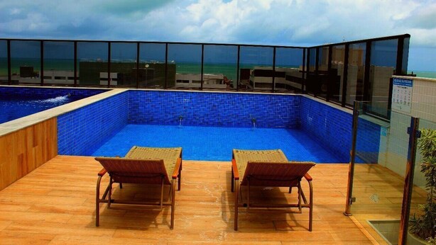 A rooftop pool with two lounge chairs, a glass balustrade, and a view of the ocean. - Compact Flat Tambau (João Pessoa)