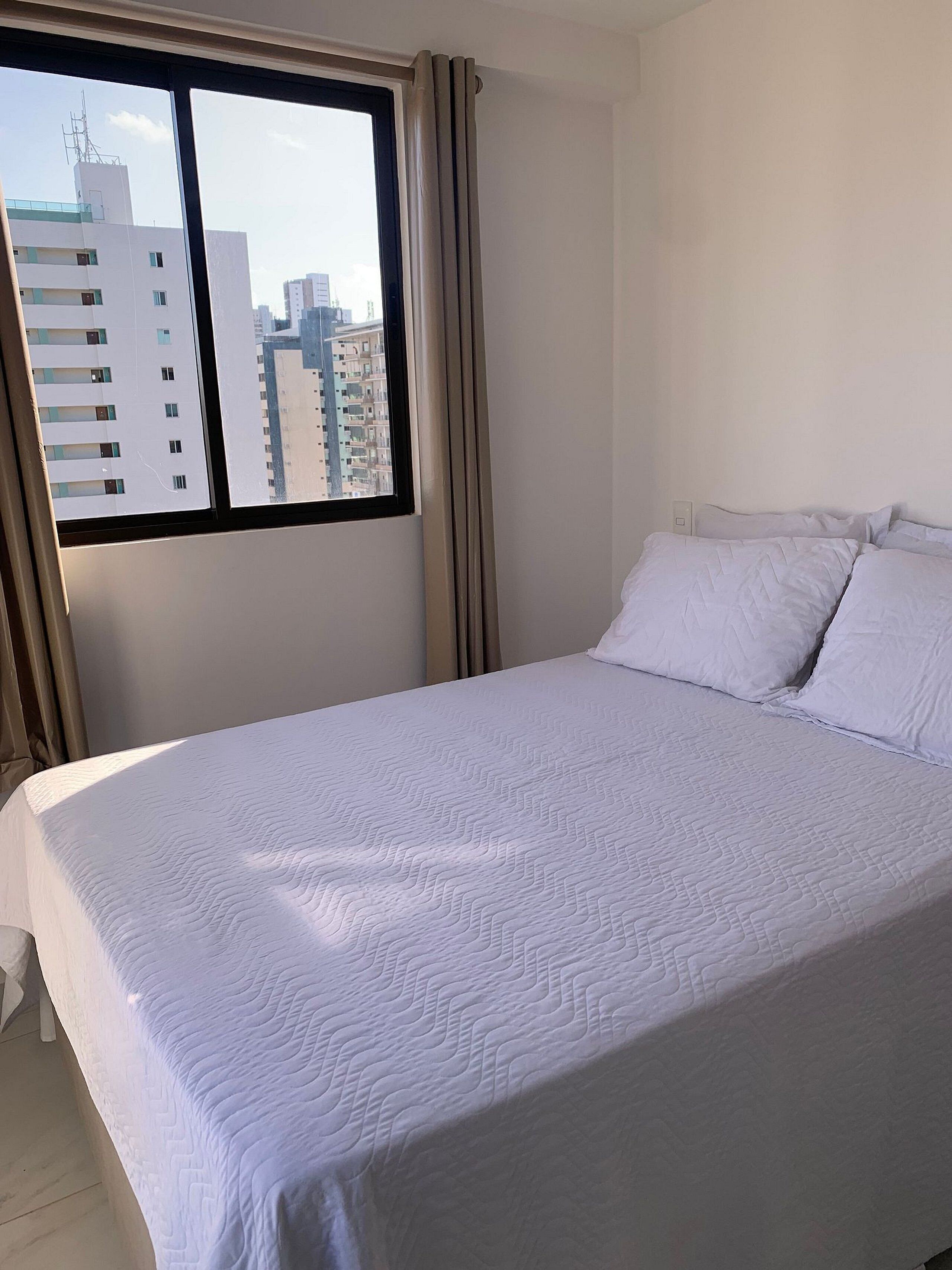 Una cama bien hecha con ropa blanca en una habitación con un ventanal grande, cortinas y vistas a edificios.