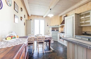 Een keuken met een eettafel, stoelen en een fruitmand. Er is een raam met gordijnen en een deur naar een andere kamer.