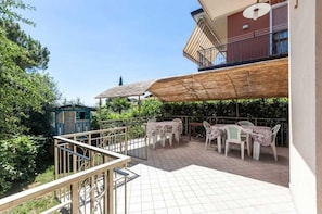 Een overdekt terras buiten met tafels en stoelen, een houten pergola en zicht op groen en gebouwen in de verte.
