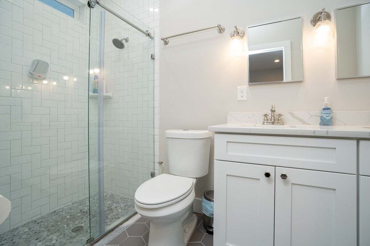 A bathroom with a glass-enclosed shower, white tiled walls, a white toilet, and a vanity with a mirror and soap dispenser.