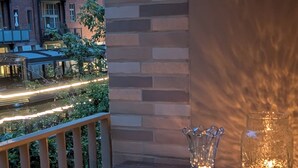 Un balcon avec une table, deux vases en verre illuminés, et une vue sur un immeuble en briques et sur les lampadaires de la rue.