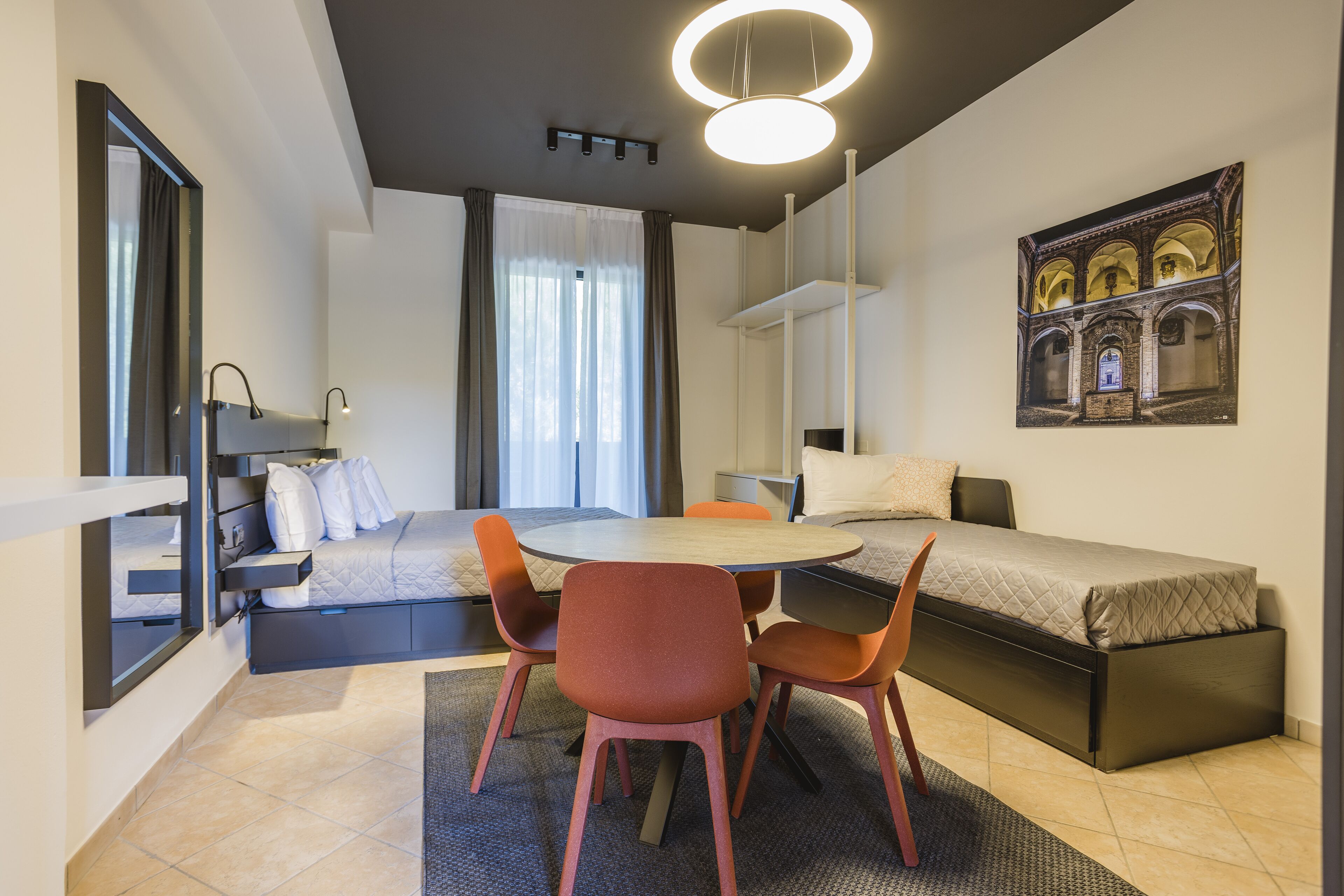 Une pièce de taille modeste comprenant un lit, une table à manger avec des chaises, un petit coin salon et une grande fenêtre avec des rideaux.