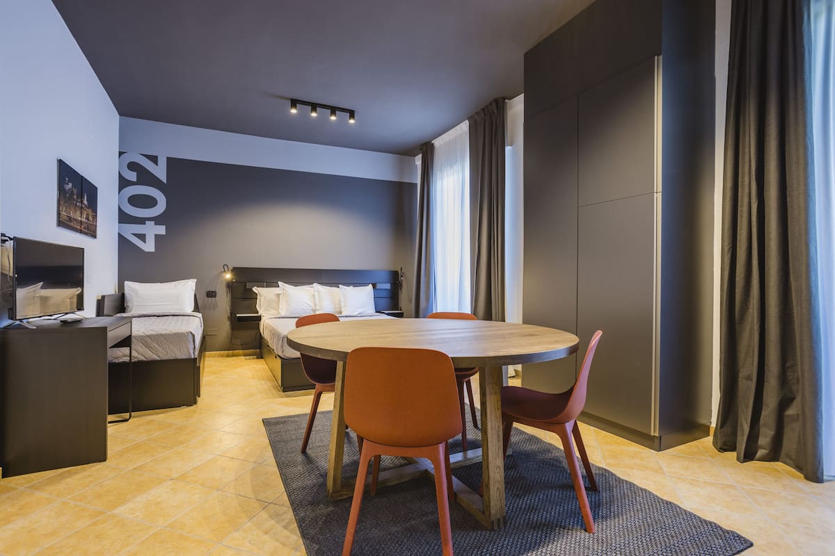 A modern hotel room with a bed, a desk, a dining table with chairs, and a large window with curtains.