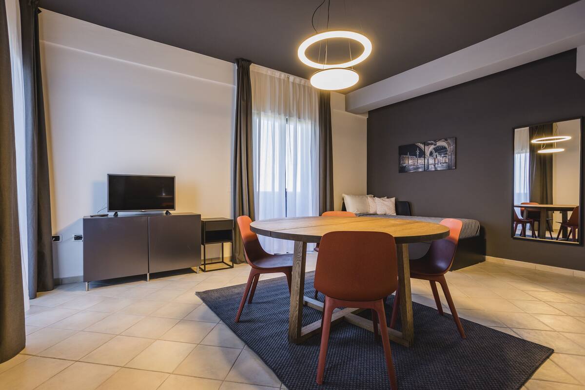 A modern hotel room with a round dining table, two chairs, a TV on a cabinet, a bed with a headboard, a painting on the wall, and a large mirror.