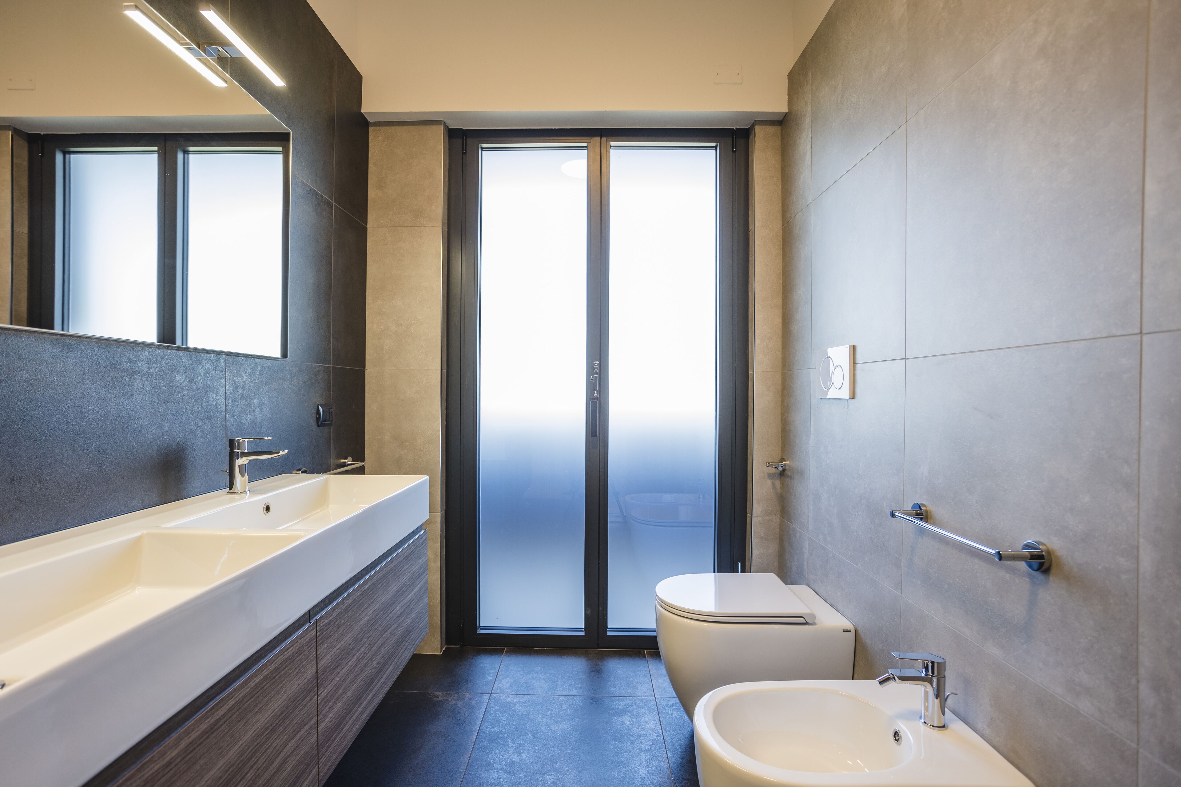Ein modernes Badezimmer mit einem großen Spiegel, einem Doppelwaschbecken, einer Toilette und einer Glastür zum Balkon.