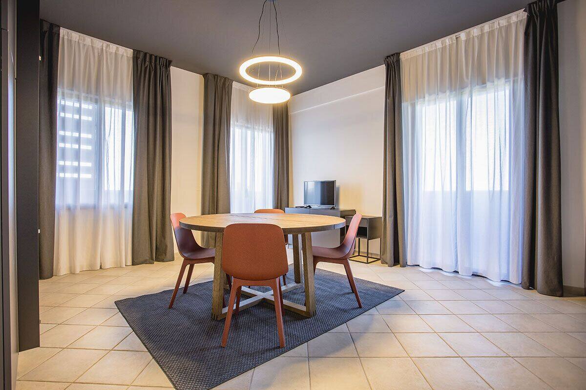 A modern dining area with a round table, four chairs, a flat-screen TV, and large windows with curtains.