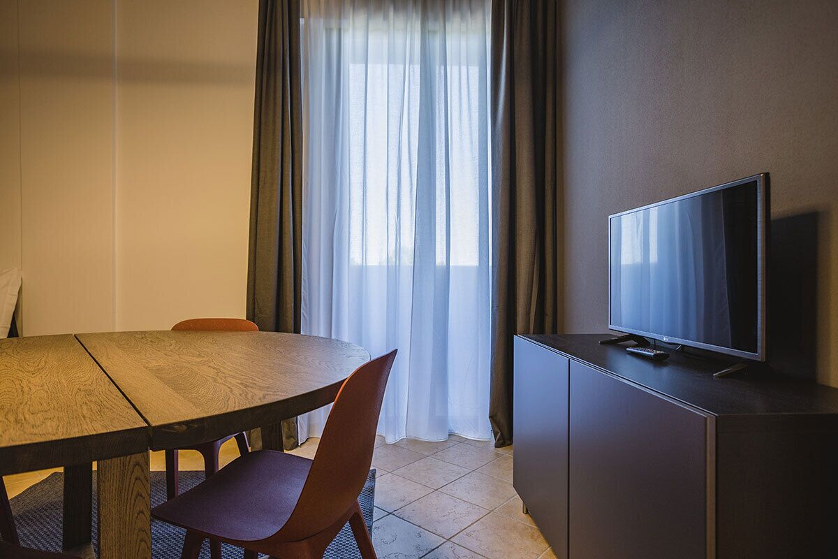 A hotel room with a wooden dining table, two chairs, a television on a cabinet, and a window with sheer curtains.