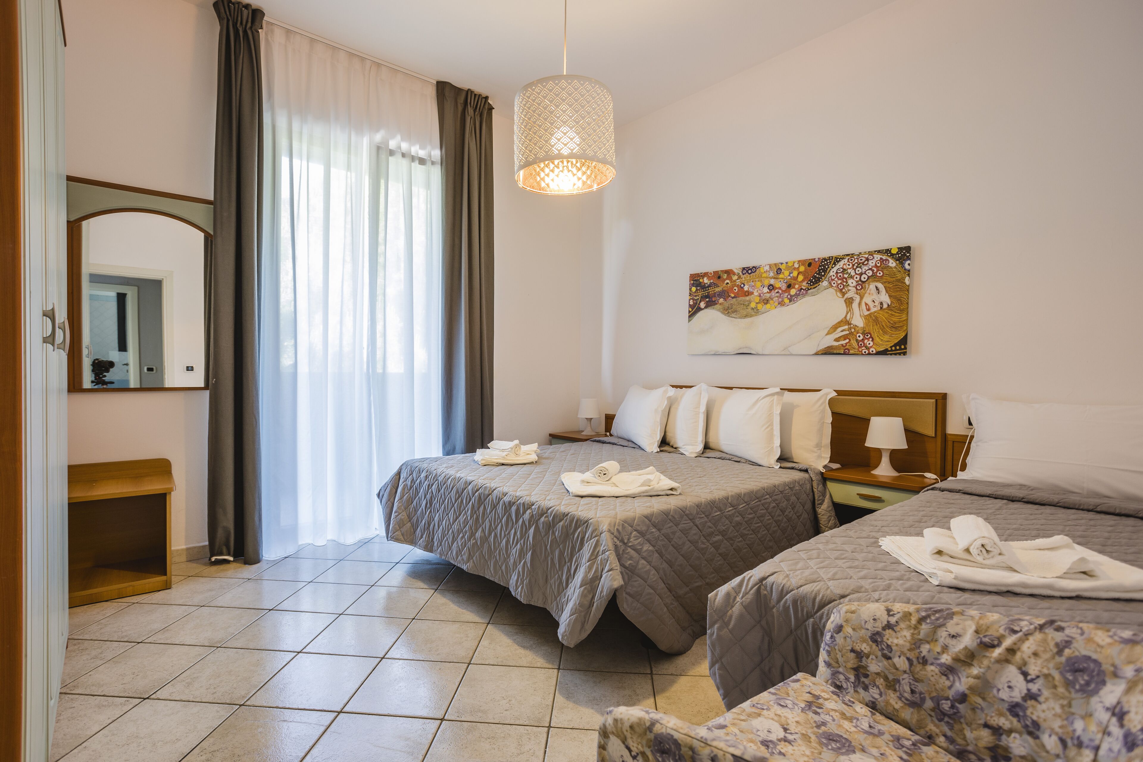 A hotel room with a large bed, a bedside table with a lamp, a painting on the wall, and a window with curtains.