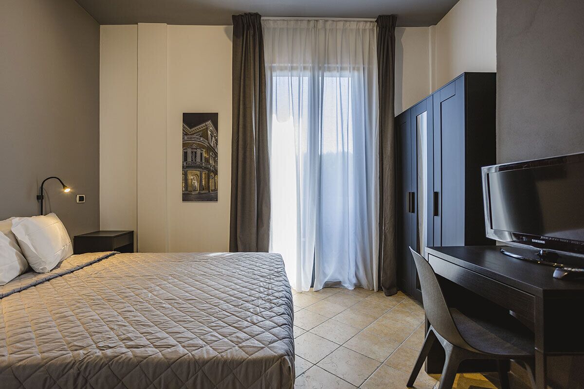 Habitación de hotel con cama, escritorio con televisor, mesita de noche y ventana con cortinas.