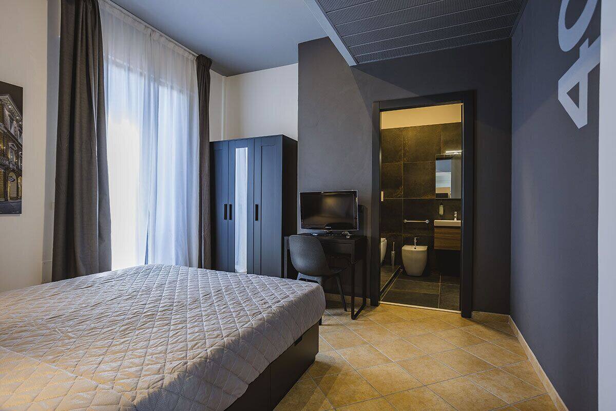 A modern hotel room with a bed, a desk with a chair, a television, and a bathroom visible through an open door.