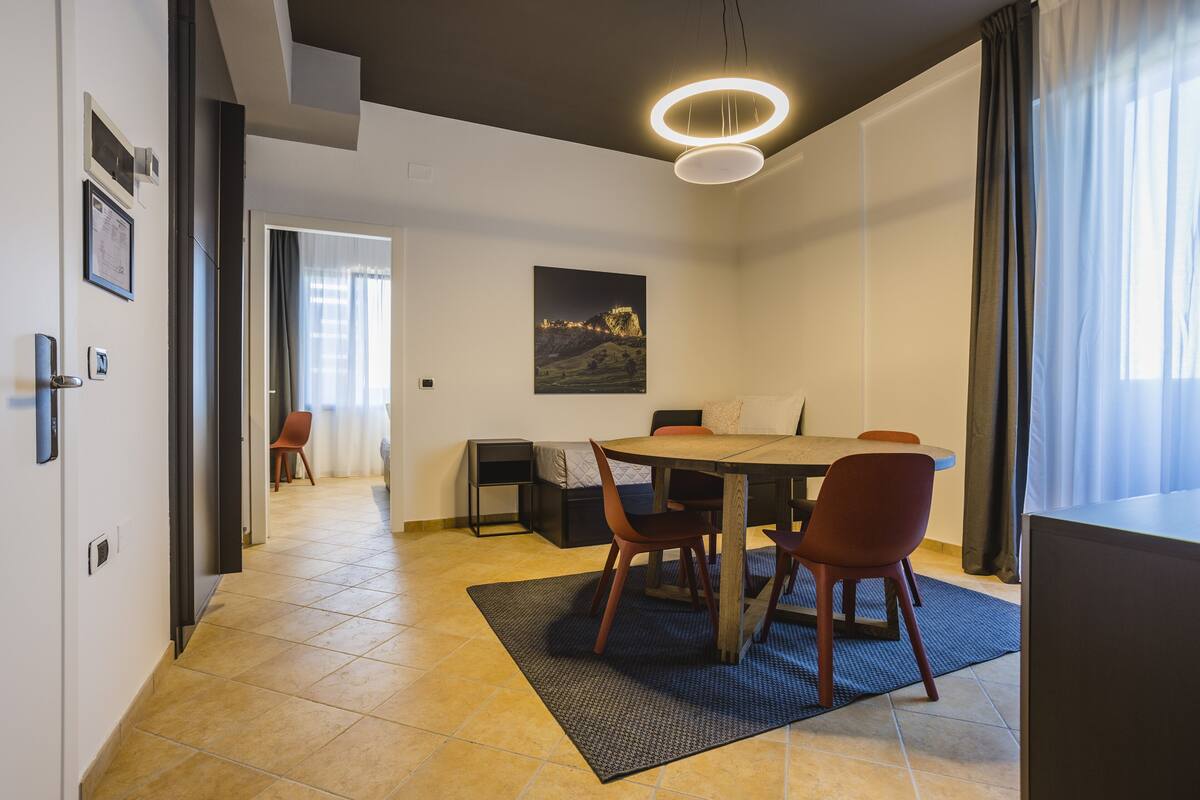 A modern dining area with a round table, chairs, and a pendant light. There is a painting on the wall and a window with curtains.