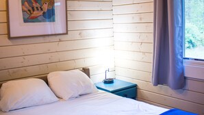 A single bed with white bedding and a blue blanket, a bedside table with a lamp, a framed picture on the wall, and a window with curtains. - Carowinds Camp Wilderness (Charlotte)