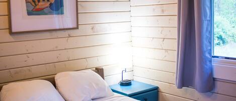 Un lit simple avec une literie blanche et une couverture bleue, une table de chevet avec une lampe, un tableau encadré au mur et une fenêtre avec des rideaux.