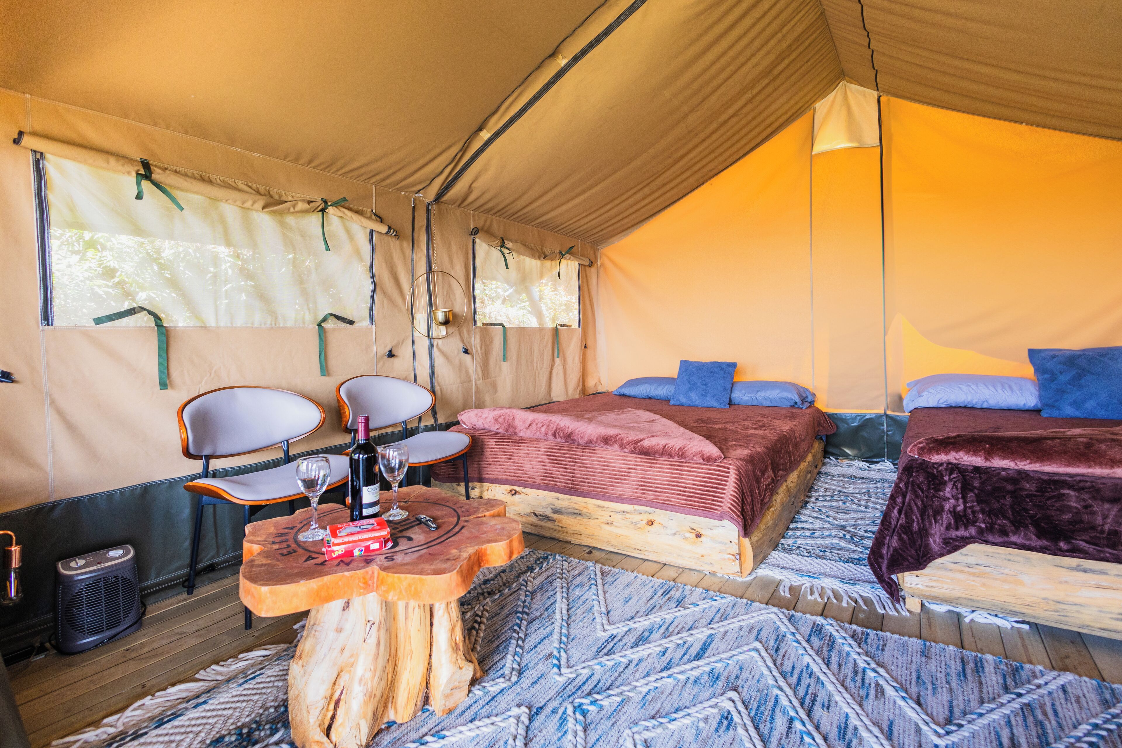 A cozy tented accommodation with a bed, two chairs, a small table with a bottle and glass, and a woven rug.