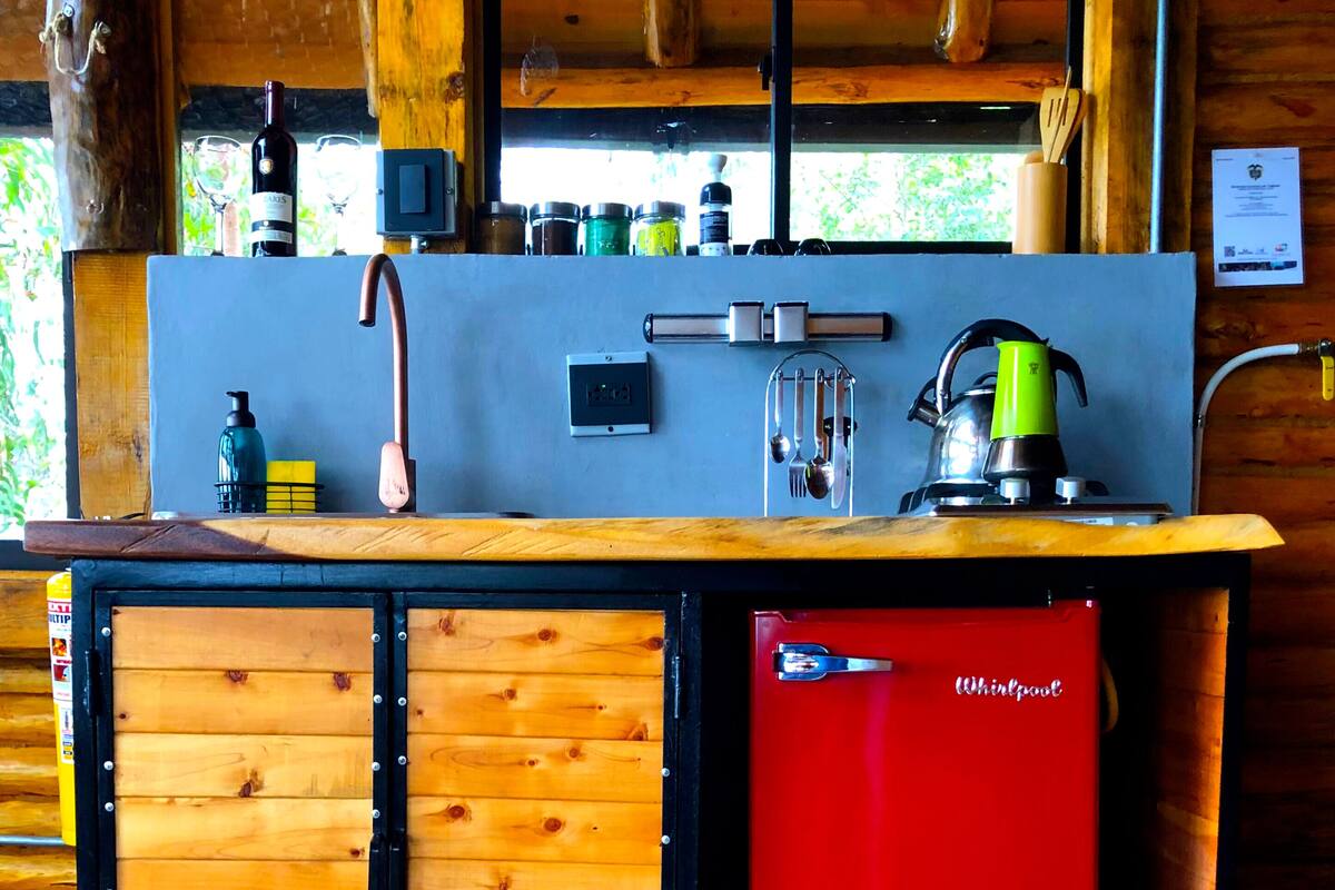A wooden cabin interior with a kitchen area featuring a sink, a red Whirlpool refrigerator, and a window with a view of greenery.