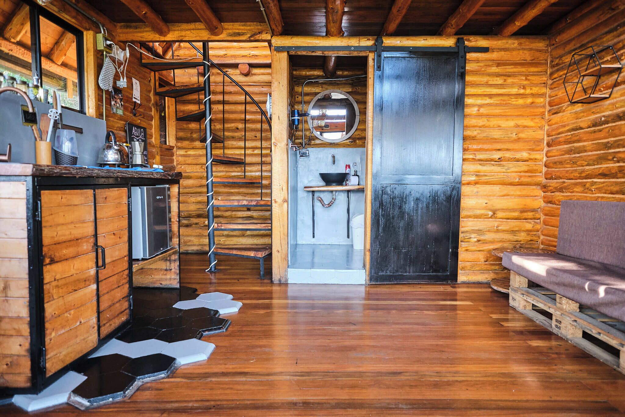 Interior rústico de una cabaña con paredes de madera, una cocina con pileta y tetera, y una escalera de caracol.