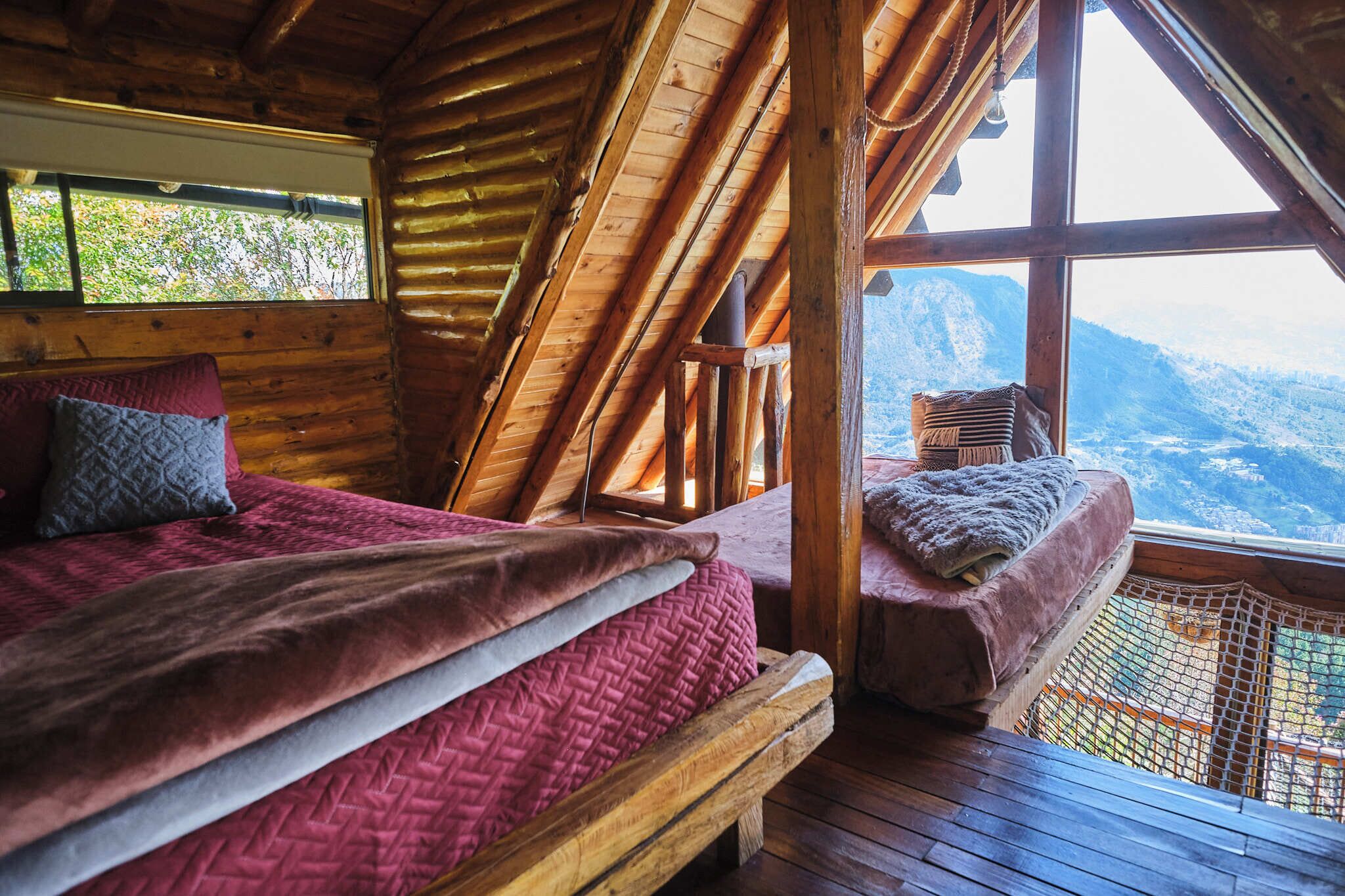 A wooden cabin with a loft-style bedroom featuring two beds, a large window, and a view of the mountains.