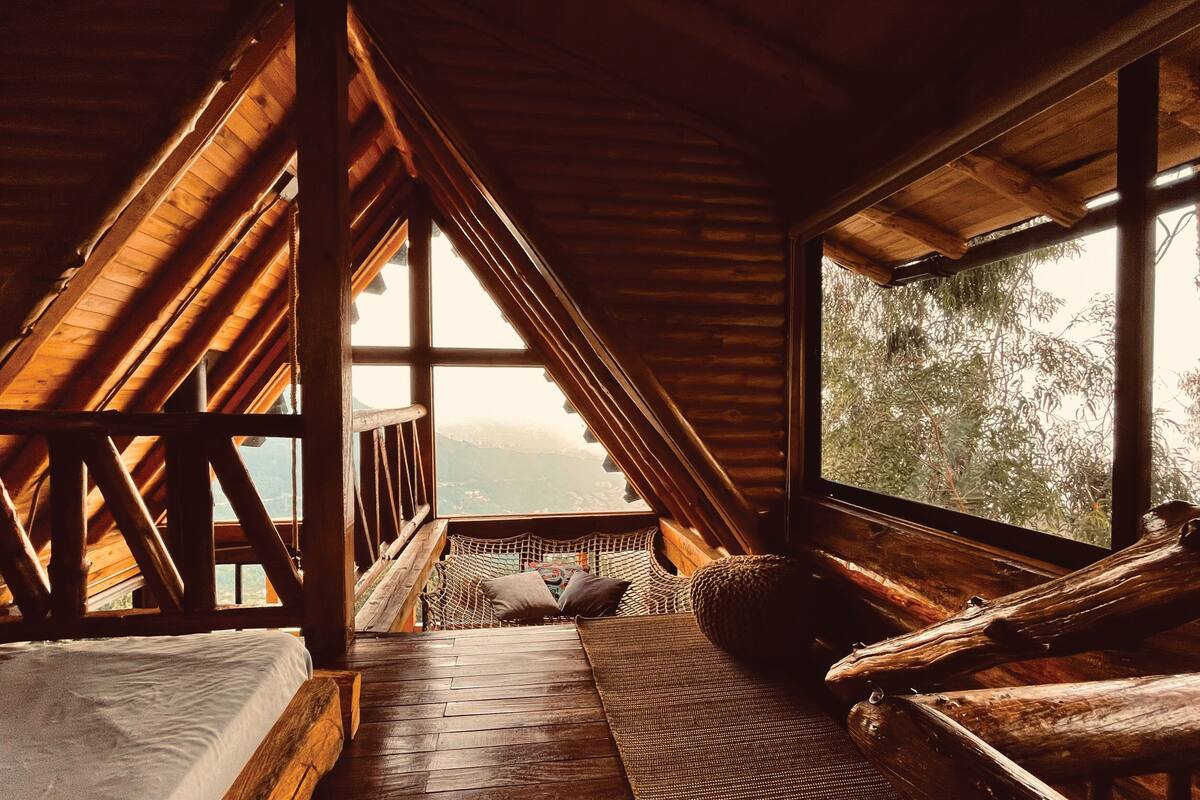 A high-ceilinged room with a sloped roof, a large window offering a view of the outdoors, and a cozy seating area with a bed and cushions.