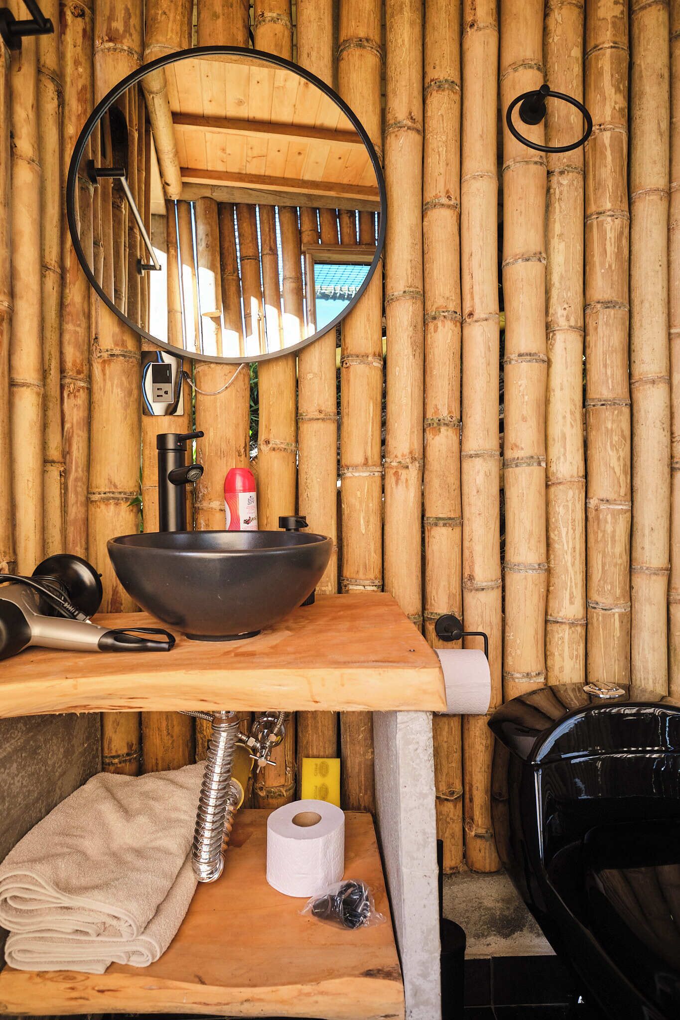 A bathroom with a bamboo wall, a round mirror, a black sink, and a towel on the counter.