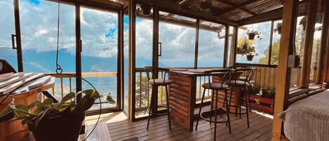 A wooden deck with a table and chairs, large windows with a view of the sea, and hanging plants.
