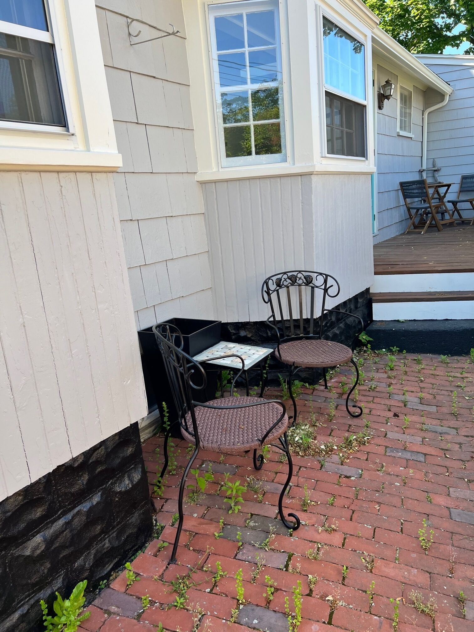 Une terrasse avec deux fauteuils en osier et une petite table, un chemin en briques et un bâtiment blanc doté de fenêtres.