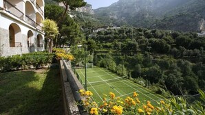 A tennis court with a view of a mountainous landscape, a building with arches, and a garden with yellow flowers. - Splendid Suite with Sea View Terrace  (Positano)