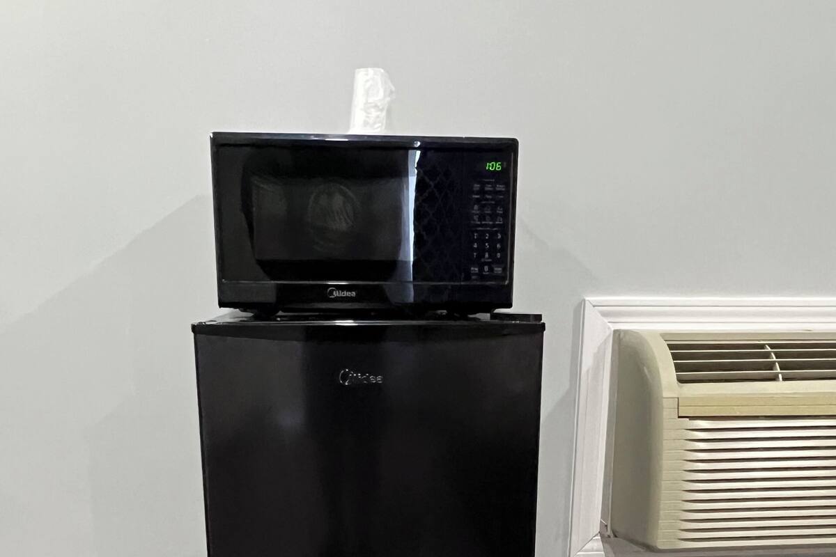 Uma geladeira preta com um micro-ondas em cima, uma parede branca e uma janela com persianas.