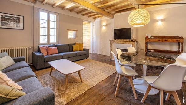 A living room with a sofa, coffee table, and dining table with chairs. There is a fireplace, a television, and a window with blinds. - Maison 116 - Hypercentre - Gaillac (Gaillac)