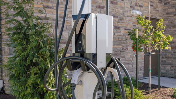 Une borne de recharge pour véhicules électriques dotée de plusieurs câbles, un bâtiment en briques en arrière-plan et un petit arbre à proximité.