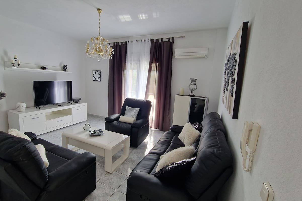 A modern living room with a flat-screen TV, leather sofas, a coffee table, and a chandelier.