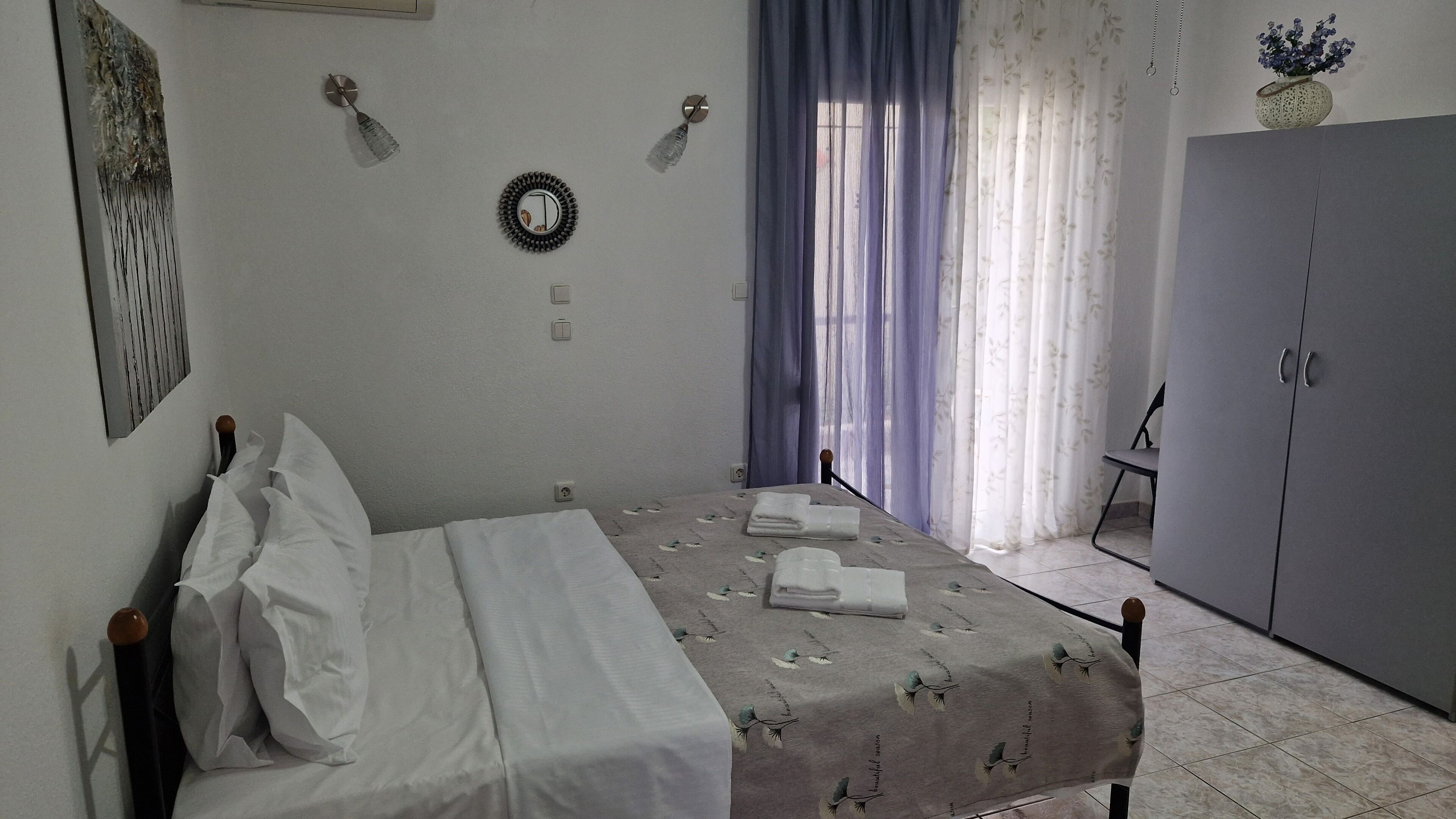 Un dormitorio con cama, armario, espejo en la pared y ventana con cortinas.