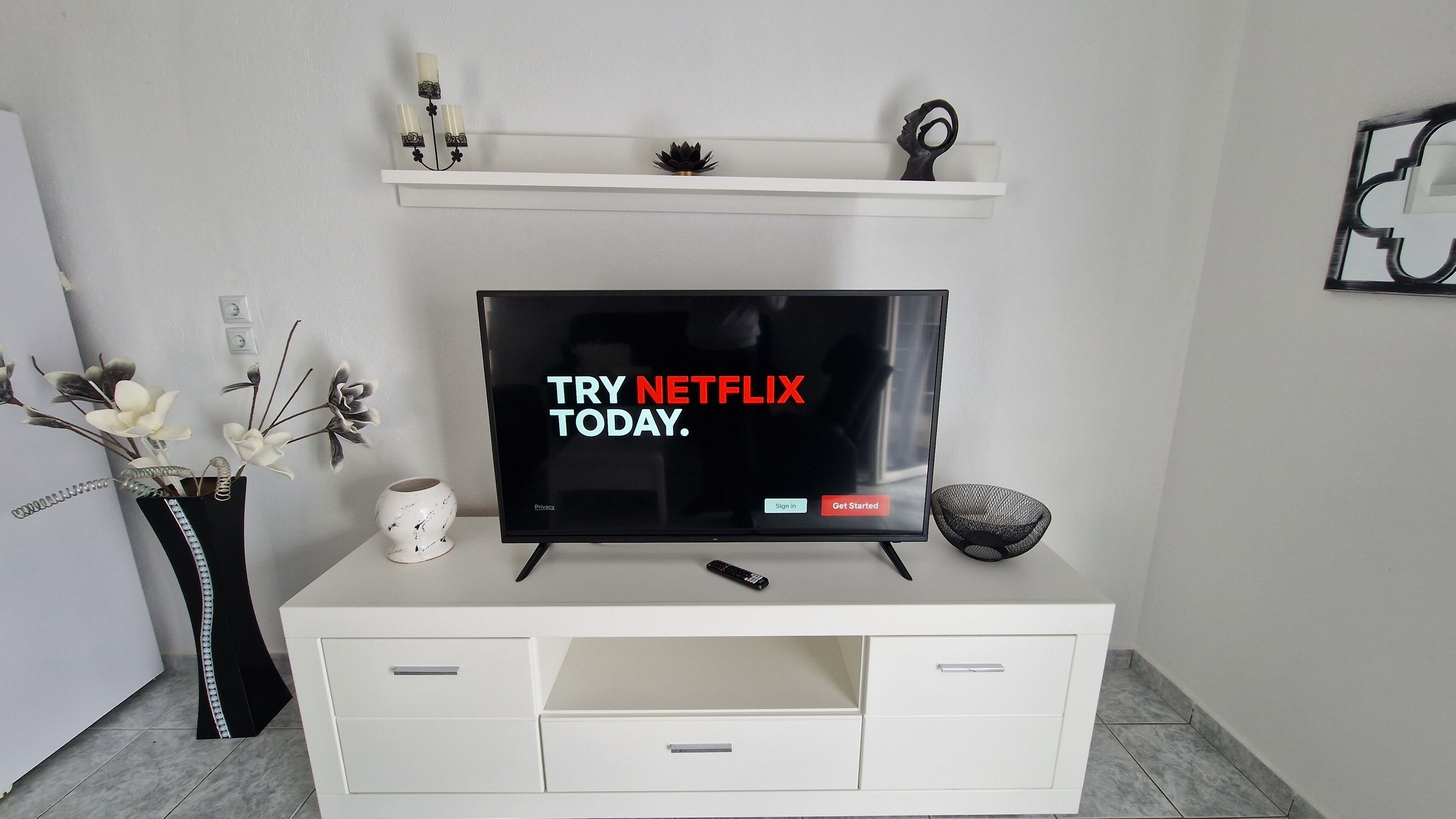 Una sala de estar moderna con un televisor de pantalla plana mostrando el logotipo de Netflix, un mueble blanco con cajones, un jarrón con flores y un estante con objetos decorativos.