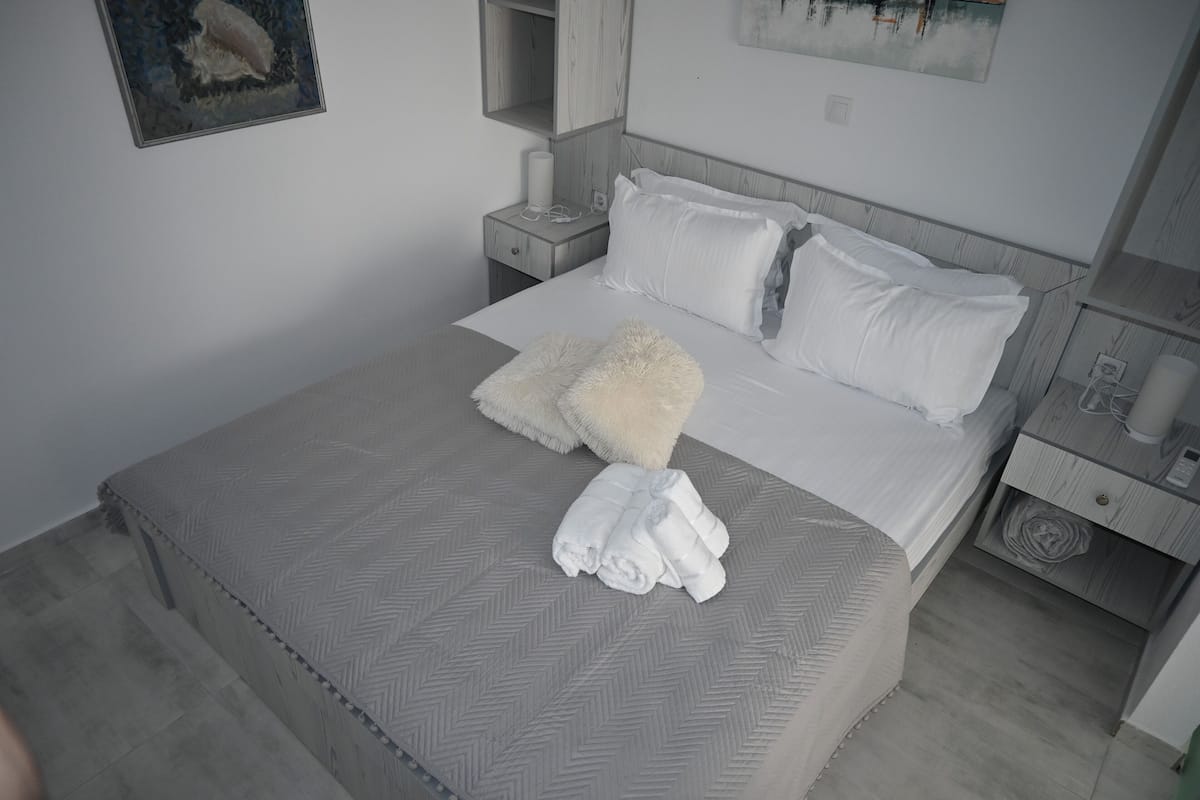 A neatly made bed with white linens, a fluffy white throw blanket, and rolled towels on a grey herringbone-patterned bedspread.