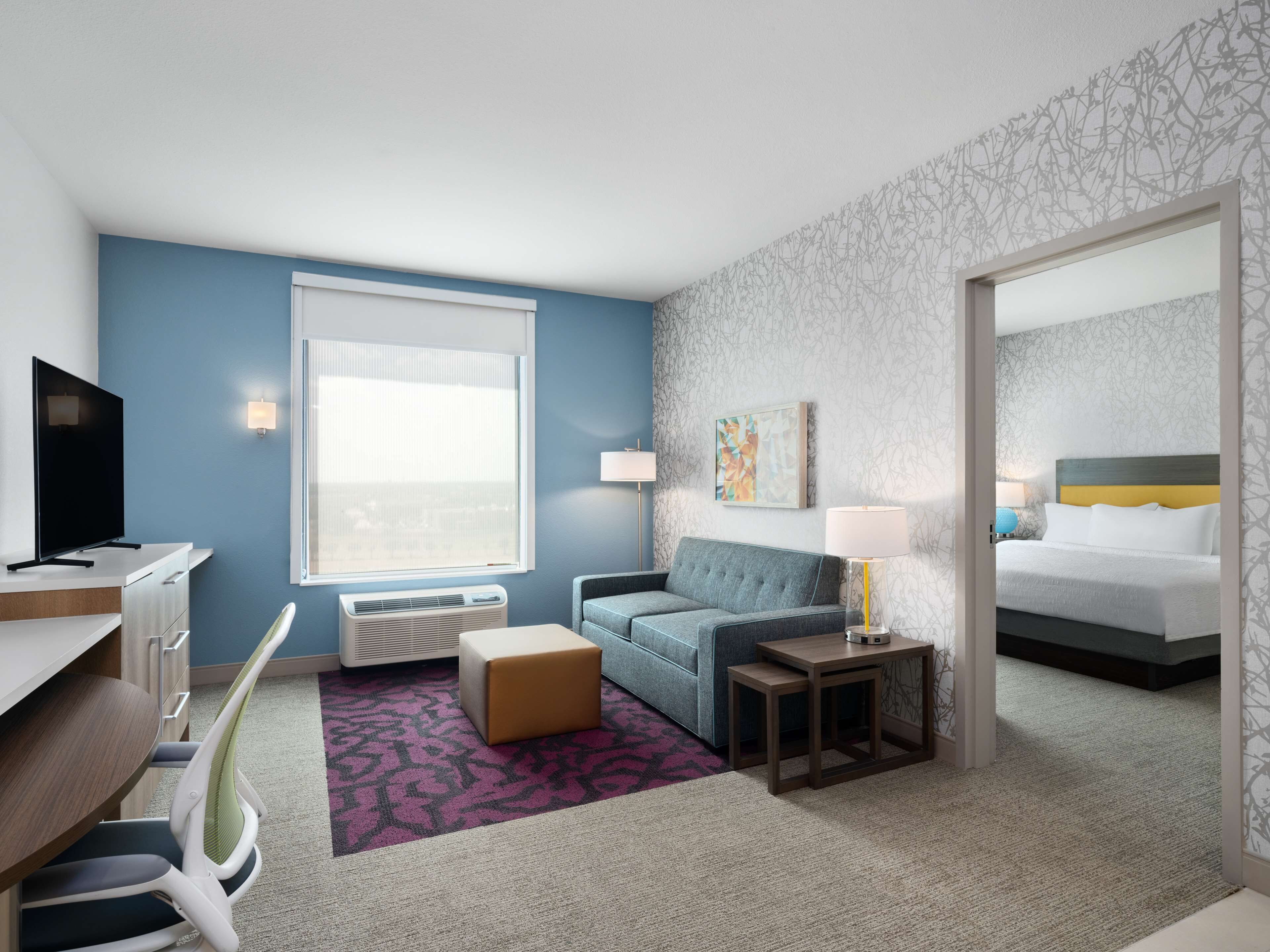 A hotel room with a desk, chair, sofa, bedside table, lamp, and a bed.