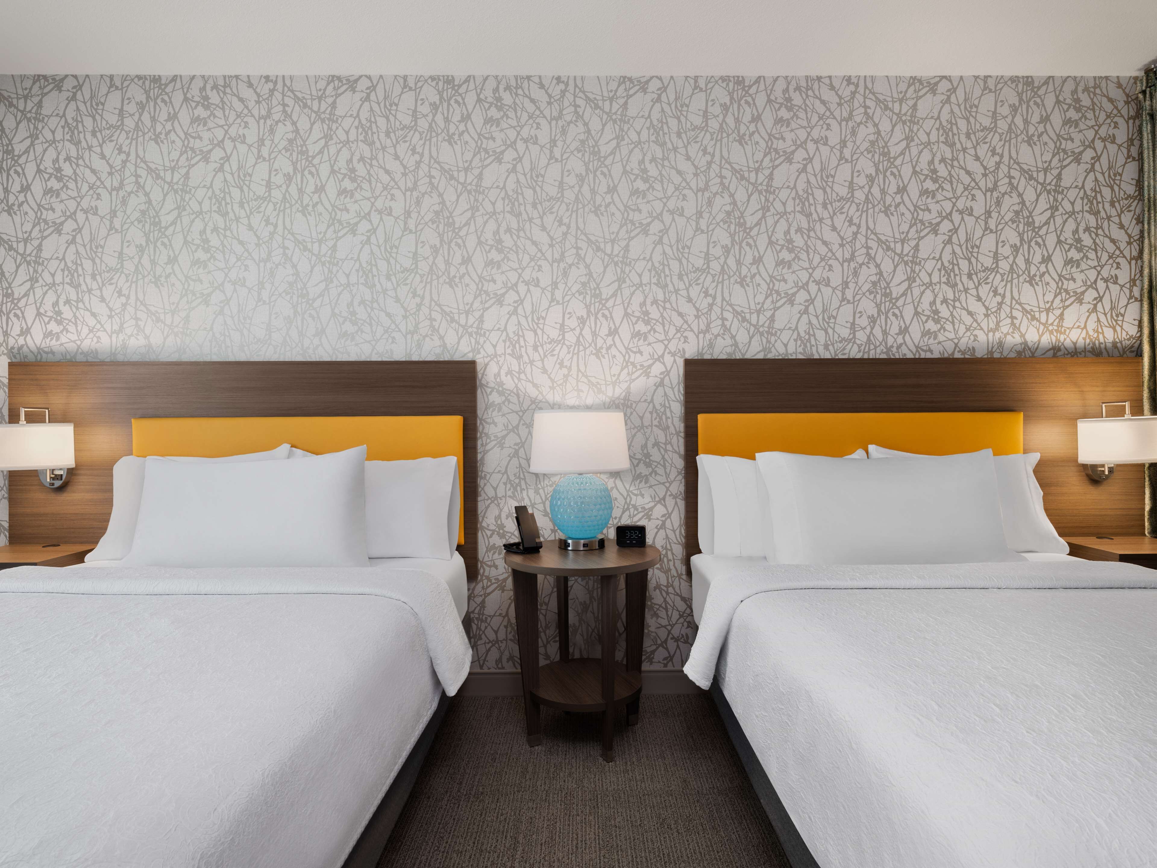 Habitación de hotel con dos camas, cada una con cabecero de madera y ropa de cama blanca. Entre las camas, una pequeña mesa con una lámpara y un globo decorativo.